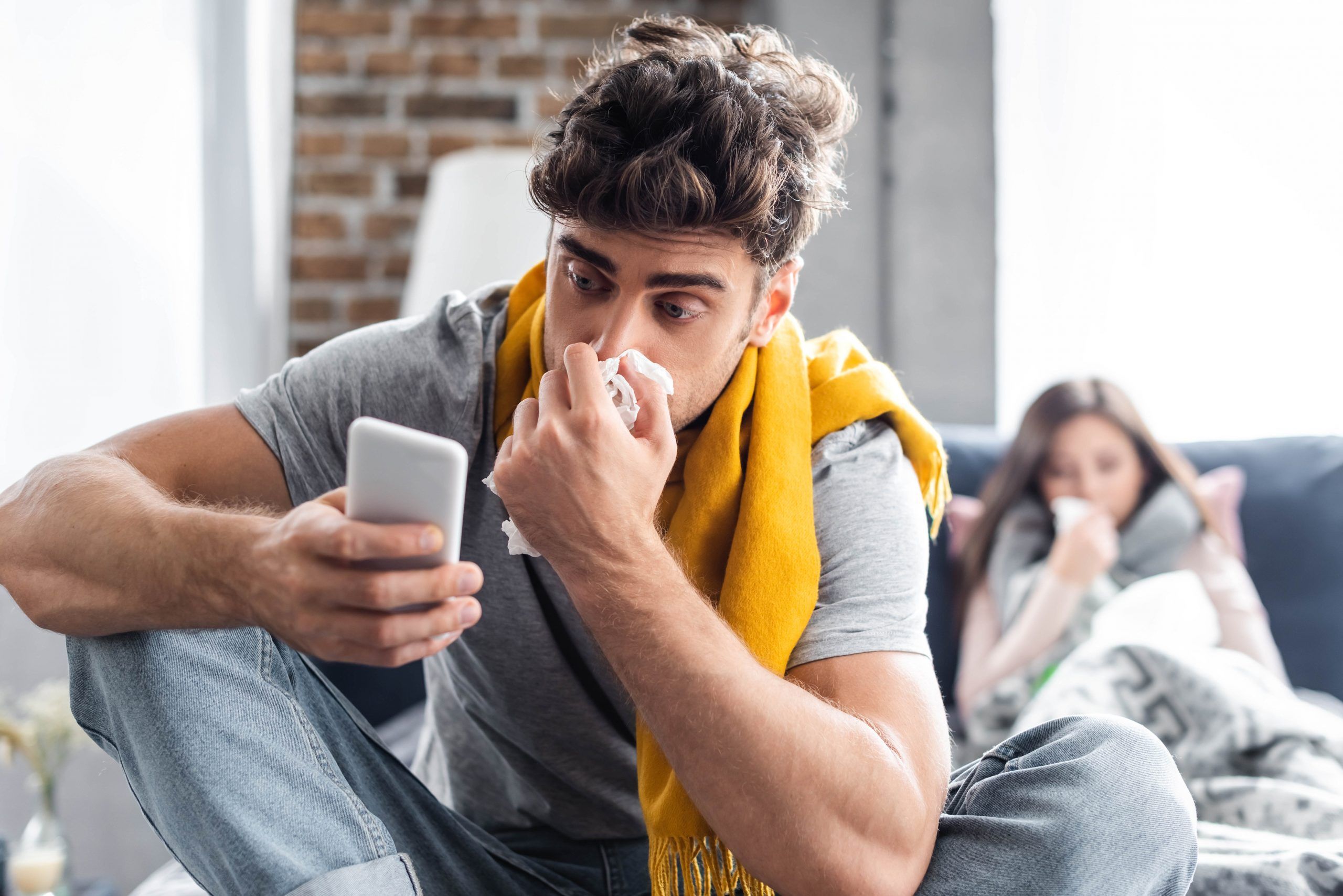 Mann putzt sich die Nase und schaut in sein Handy. Frau sitzt auf Sofa und hält ein Taschentuch